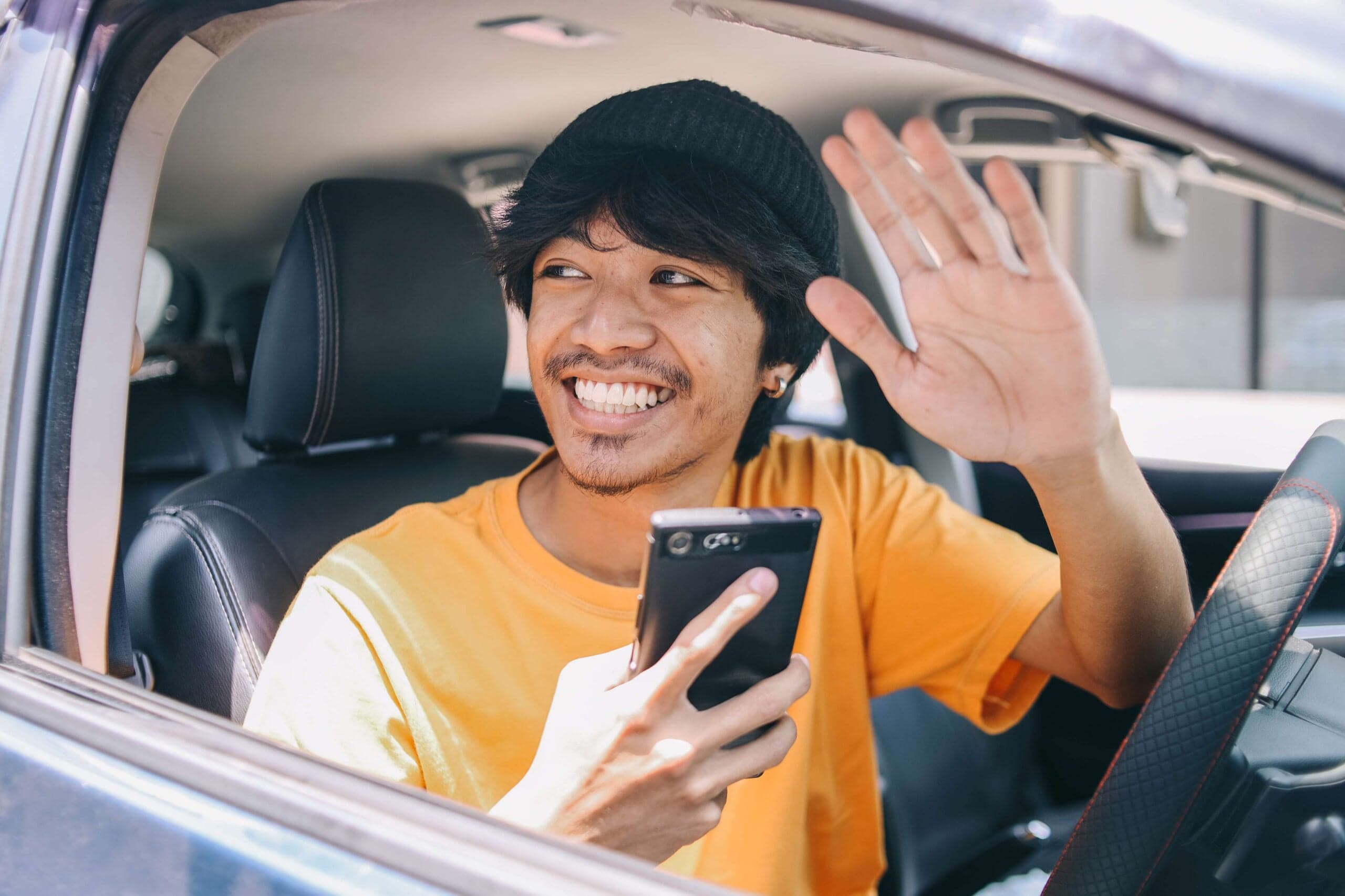 A smiling driver waves while holding his smartphone in the car